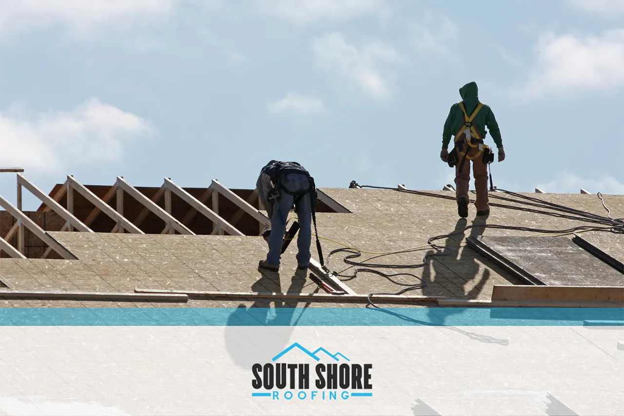 Two workers on a rooftop in Savannah are installing roofing materials with tools in hand, wearing safety harnesses. The logo "South Shore Roofing" is displayed at the bottom of the image, highlighting their expertise in winter roof replacement.